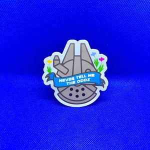 Può includere: Un adesivo raffigurante il Millennium Falcon con la scritta "NEVER TELL ME THE ODDS" su uno striscione blu. L'adesivo ha un bordo bianco ed è decorato con piccoli fiori.