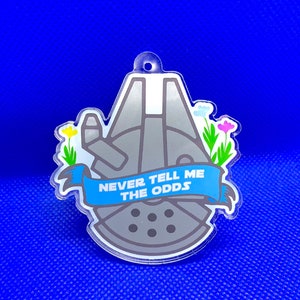 Può includere: Un ciondolo in acrilico trasparente con un design grigio del Millennium Falcon e uno striscione blu con la scritta "NEVER TELL ME THE ODDS". Il ciondolo è decorato con fiori colorati e ha un piccolo foro per appenderlo.