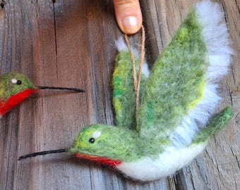 May include: Two needle felted hummingbird ornaments. One is green with white and yellow accents and a red throat. The other is green with a red throat. Both have black eyes and beaks.