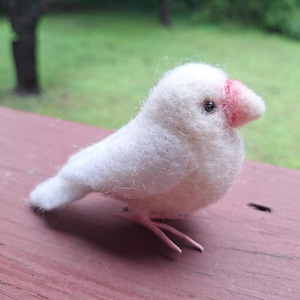 May include: A handmade white felt bird with a pink beak and feet. The bird is perched on a wooden surface, with a blurred green background. The bird's eye is a small black bead.