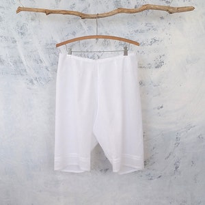 May include: White cotton undergarment with lace trim hanging on a wooden hanger against a textured white background.