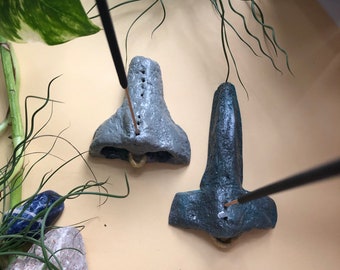 May include: Two gray and teal incense holders shaped like noses, with holes for incense sticks. The holders have a textured, stone-like appearance and small gold-colored accents. Incense sticks are inserted into the holes.