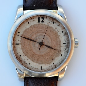 May include: A wristwatch with a silver-colored case and a brown leather band. The watch face features a natural wood grain design with a dark brown hour and minute hands. The number 12 is visible at the top of the watch face.