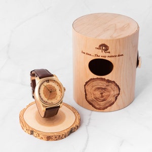 K&ouml;nnte beinhalten: Eine goldfarbene Armbanduhr mit braunem Lederarmband und einem Zifferblatt aus Holz. Die Uhr wird auf einem Holzst&auml;nder pr&auml;sentiert und kommt in einer Holzkiste mit dem Text "Tree Ring" und "See time... The way nature does." Die Box ist aus hellem Holz mit einem dunklen Holz-Baumring-Design.