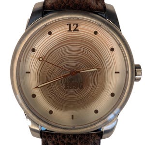 May include: A wristwatch with a brown textured leather strap and a silver-toned metal case. The watch face features a light brown wood grain pattern with concentric rings, the number 12 at the top, the year 1996 at the bottom, and thin hands.