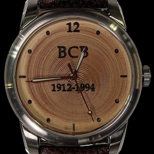 May include: A silver wristwatch with a brown leather band. The watch face has a wood grain design with the text "BCB 1912-1994" and the number "12" at the top.