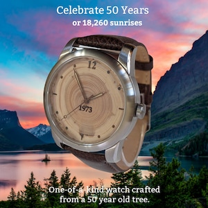 May include: A brown leather wristwatch with a silver metal case. The watch face is made of wood with a tree ring pattern and the numbers 12 and 1973. The text "Celebrate 50 Years or 18,260 sunrises" is at the top of the image. The watch is in front of a mountain range and a lake.