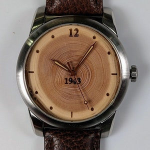 K&ouml;nnte beinhalten: Eine silberne Armbanduhr mit einem braunen Lederarmband. Das Zifferblatt der Uhr hat ein Holzmaserungsmuster mit der Zahl 12 und dem Jahr 1943.