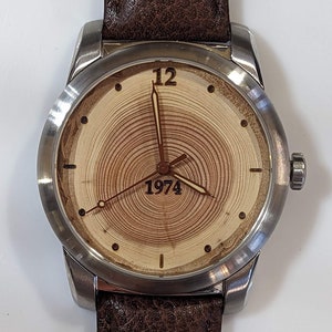 May include: A silver wristwatch with a brown leather band. The watch face has a wood grain design with the number 12 and the year 1974.