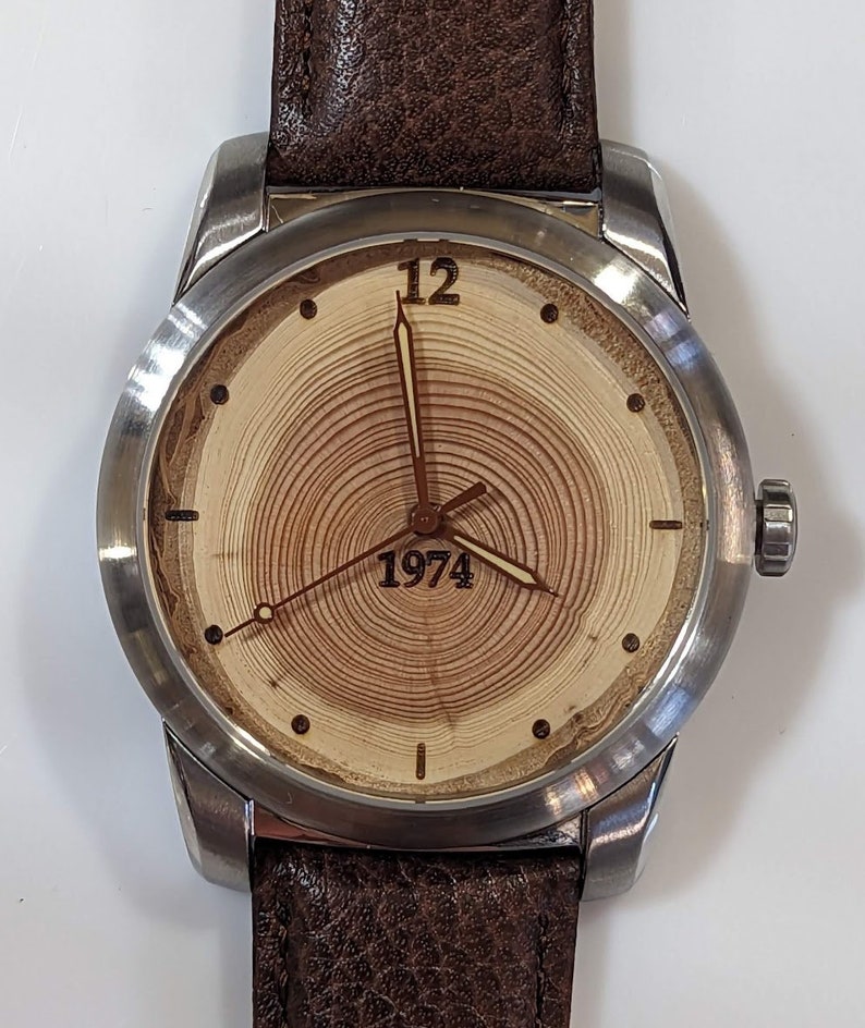 May include: A silver wristwatch with a brown leather band. The watch face has a wood grain design with the number 12 and the year 1974.