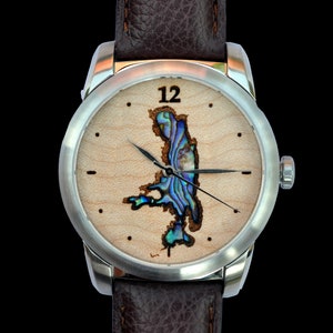 Puede incluir: Un reloj de pulsera de acero inoxidable con una correa de cuero marrón. La esfera del reloj presenta un fondo de veta de madera clara con una incrustación colorida similar a la del abulón. Las manecillas del reloj son negras y el número 12 se muestra en la parte superior.