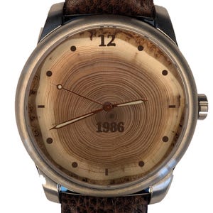 May include: A wristwatch with a silver-toned metal case and a dark brown textured leather strap. The watch face features a natural wood grain pattern resembling tree rings, with the number 12 at the top and the year 1986 printed in the center.