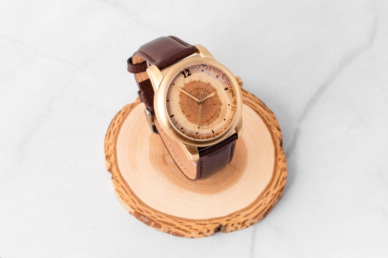 May include: A gold-tone wristwatch with a brown leather band. The watch face has a unique wood grain design with a gold-tone hour and minute hand.