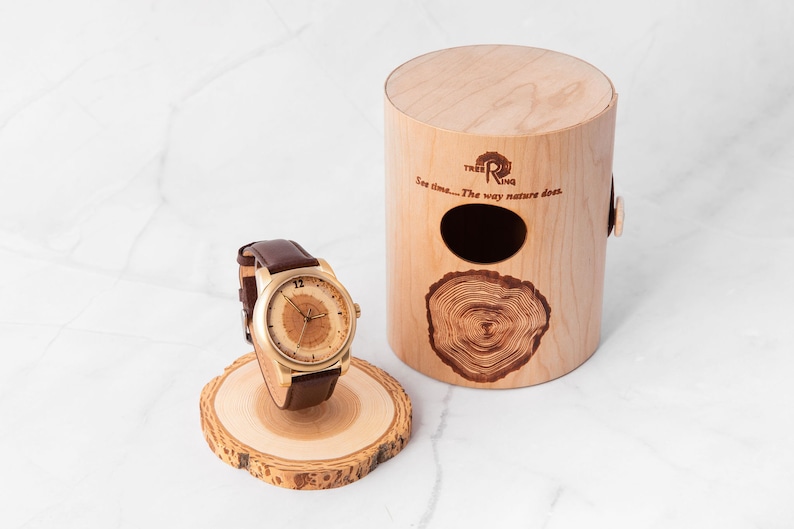 May include: A gold-tone wristwatch with a brown leather band and a wooden face. The watch is displayed on a wooden stand and comes in a wooden box with the text "Tree Ring" and "See time... The way nature does." The box is made of light wood with a dark wood tree ring design.
