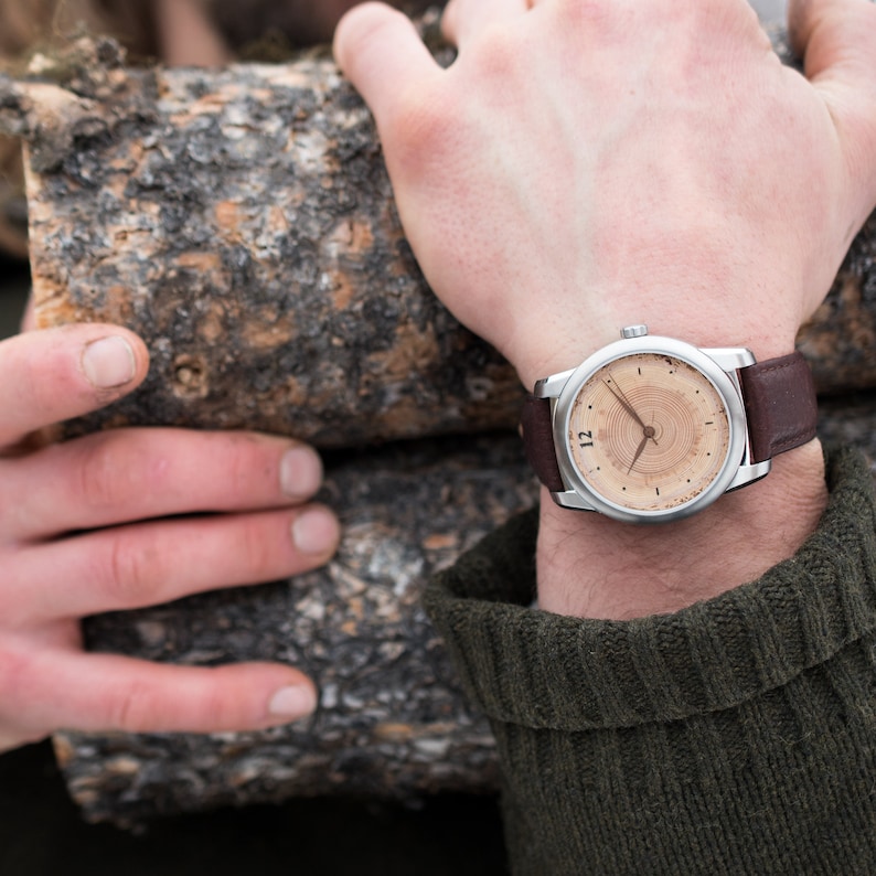 K&ouml;nnte beinhalten: Eine silberne Armbanduhr mit einem braunen Lederarmband. Das Zifferblatt der Uhr hat ein Holzmaserungsmuster mit einem kreisf&ouml;rmigen Muster und die Zahl 12.