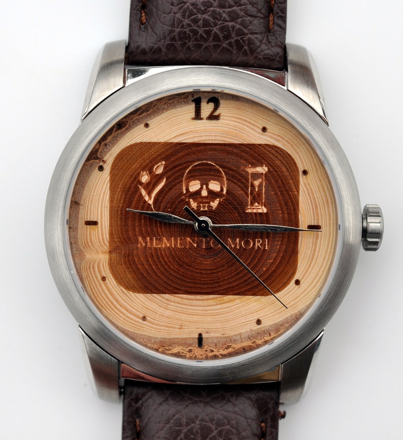 May include: A silver wristwatch with a brown leather strap. The watch face is made of wood with a skull, flower, and hourglass design. The text "Memento Mori" is engraved on the wood.