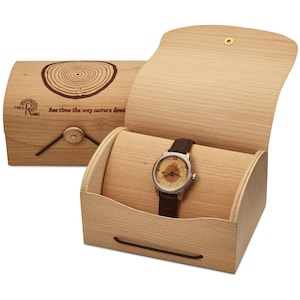 May include: A wooden watch box with a brown leather watch inside. The box is made of light wood and has a hinged lid. The watch has a round face with a tree ring design and the words "See time the way nature does" on the lid.