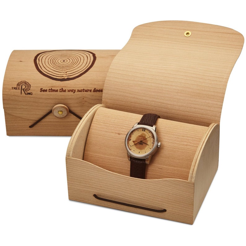 May include: A wooden watch box with a brown leather watch inside. The box is made of light wood and has a hinged lid. The watch has a round face with a tree ring design and the words "See time the way nature does" on the lid.