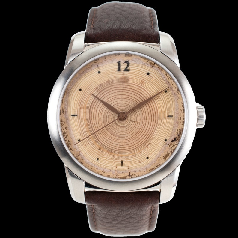 May include: A silver wristwatch with a brown leather strap. The watch face has a wood grain design with a circular pattern and black hour and minute hands. The number 12 is printed in black at the top of the watch face.