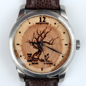 May include: A silver and wood wristwatch with a brown leather strap. The watch face features a tree with the words "Memento Mori Vivere" and "12 Memento" printed on the face.