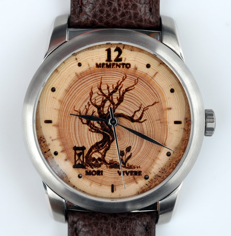 May include: A silver and wood wristwatch with a brown leather strap. The watch face features a tree with the words "Memento Mori Vivere" and "12 Memento" printed on the face.