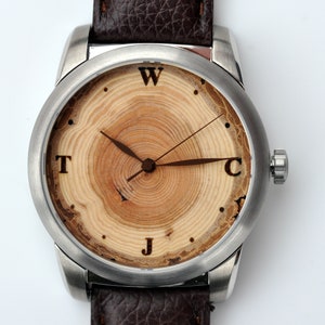 May include: A silver wristwatch with a brown leather strap. The watch face is made of wood with a tree ring pattern and has the letters W, T, J, and C around the edge.
