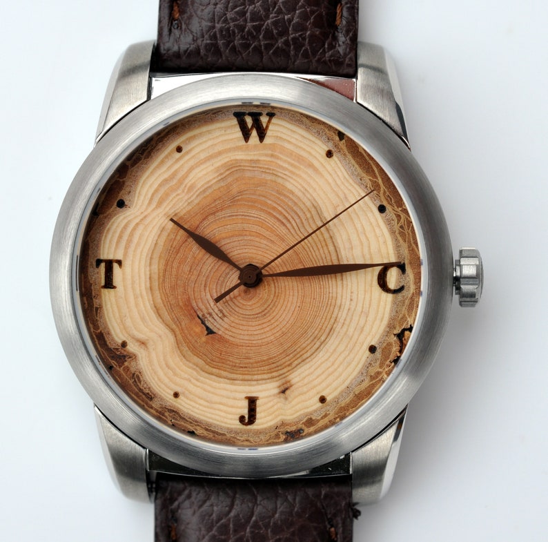 May include: A silver wristwatch with a brown leather strap. The watch face is made of wood with a tree ring pattern and has the letters W, T, J, and C around the edge.