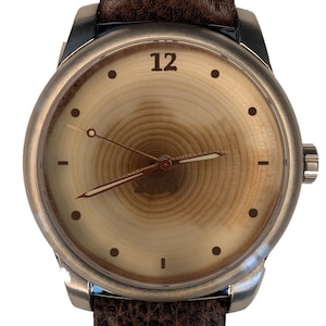 May include: A wristwatch with a round silver-tone case and a textured dark brown leather strap. The watch face features a light tan wood grain pattern with concentric rings, a single black numeral 12, black dot hour markers, and thin hands.