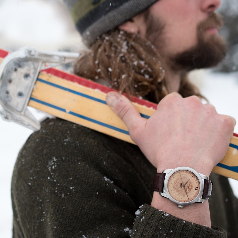 May include: A man wearing a brown sweater and a brown leather watch with a wooden face. The watch face has the number 12 and the word "TRVL" on it. The man is holding a wooden ski.