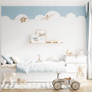 May include: A bedroom featuring a wall with a blue and white cloud mural, two hanging wooden toy aeroplanes, a white bed with blue patterned bedding, a wooden shelf with framed aeroplane art, a white storage unit, and a vintage-style toy race car.