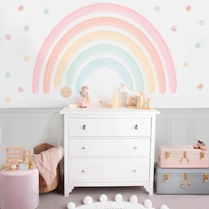 Pode incluir: Uma cômoda branca com três gavetas e puxadores dourados, em frente a uma parede com um mural de arco-íris. O quarto apresenta um pufe rosa, uma cesta de vime e itens decorativos. Um baú rosa e cinza estão empilhados.