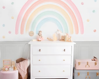 May include: A white chest of drawers with three drawers and gold knobs, set against a wall with a rainbow mural. The room features a pink ottoman, a woven basket, and decorative items. A pink and grey trunk are stacked.