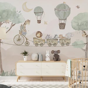 May include: A nursery room with a light brown dresser with four drawers, a white cot with a white and blue patterned blanket, and a light brown wall with a whimsical mural featuring a giraffe riding a bicycle, a lion, a zebra, and an elephant riding in a train of wagons pulled by a string. The mural also features hot air balloons and a crescent moon.