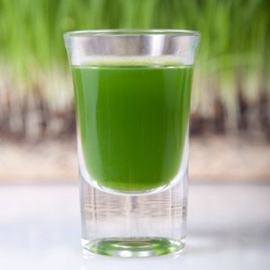 May include: A clear glass shot glass filled with bright green liquid, likely a health shot. The glass sits on a white surface, with a blurred background of green grass.