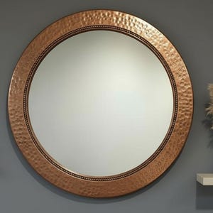 Puede incluir: Un espejo redondo grande con un marco de metal martillado de color cobre y un borde tipo cuerda. El espejo está montado en una pared gris. A la izquierda hay una obra de arte enmarcada y un candelabro, y a la derecha una estantería con un jarrón y plantas secas.