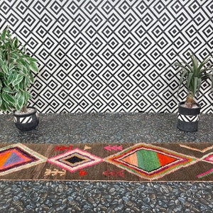 May include: A brown and black patterned rug with colorful diamond shapes. The rug is laid out on a black and white tiled floor with two potted plants on either side.