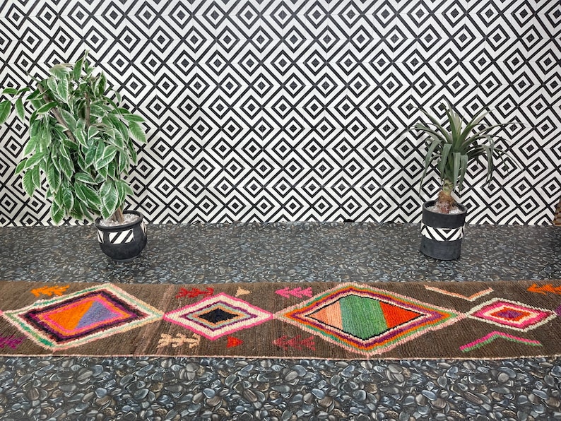 May include: A brown and black patterned rug with colorful diamond shapes. The rug is laid out on a black and white tiled floor with two potted plants on either side.