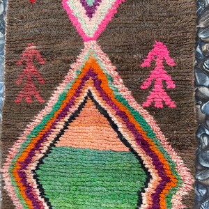 May include: A colorful Moroccan rug with a diamond pattern in the center. The rug has a brown background with pink, green, orange, and purple accents. The rug is hand-woven and has a unique, rustic look.
