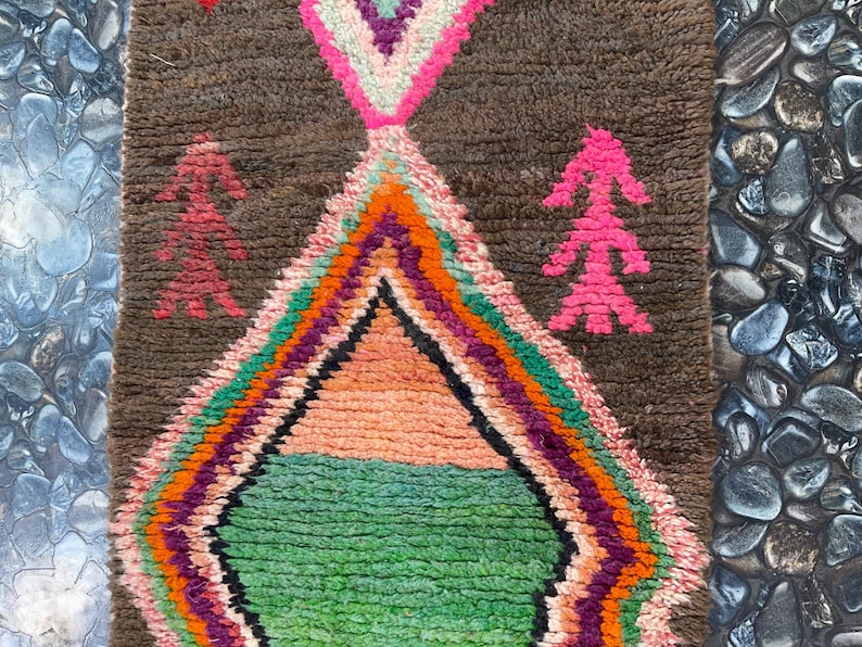 May include: A colorful Moroccan rug with a diamond pattern in the center. The rug has a brown background with pink, green, orange, and purple accents. The rug is hand-woven and has a unique, rustic look.