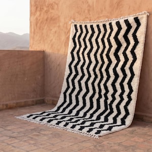 May include: A rectangular area rug featuring a high-contrast pattern of vertical black zigzag lines set against a cream-colored background. The rug has a thick, textured pile and is finished with short, knotted fringe along the top and bottom edges.