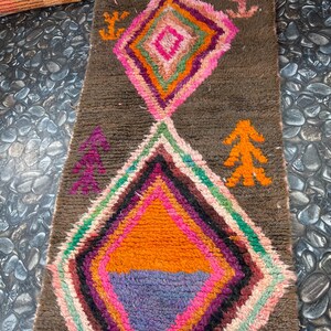 May include: A colorful Moroccan rug with a brown background and a diamond pattern. The rug has a variety of colors, including pink, orange, green, and purple. The rug is made of wool and has a soft texture.