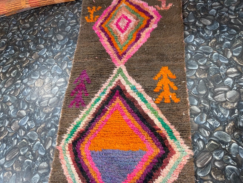 May include: A colorful Moroccan rug with a brown background and a diamond pattern. The rug has a variety of colors, including pink, orange, green, and purple. The rug is made of wool and has a soft texture.