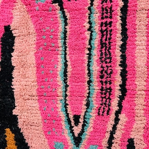 May include: Close-up of a vibrant, textured rug with a bold, colorful design. The rug features vertical stripes in shades of pink, peach, teal, and black. The design includes a central, elongated shape with a pattern and small teal dots.