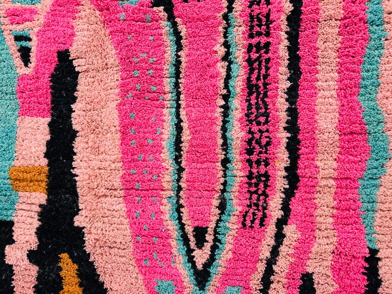 May include: Close-up of a vibrant, textured rug with a bold, colorful design. The rug features vertical stripes in shades of pink, peach, teal, and black. The design includes a central, elongated shape with a pattern and small teal dots.