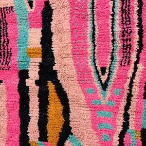 May include: Close-up of a colorful rug with a geometric pattern. The rug features a vibrant mix of colors, including pink, black, teal, and gold. The design includes vertical stripes and abstract shapes, creating a textured surface.
