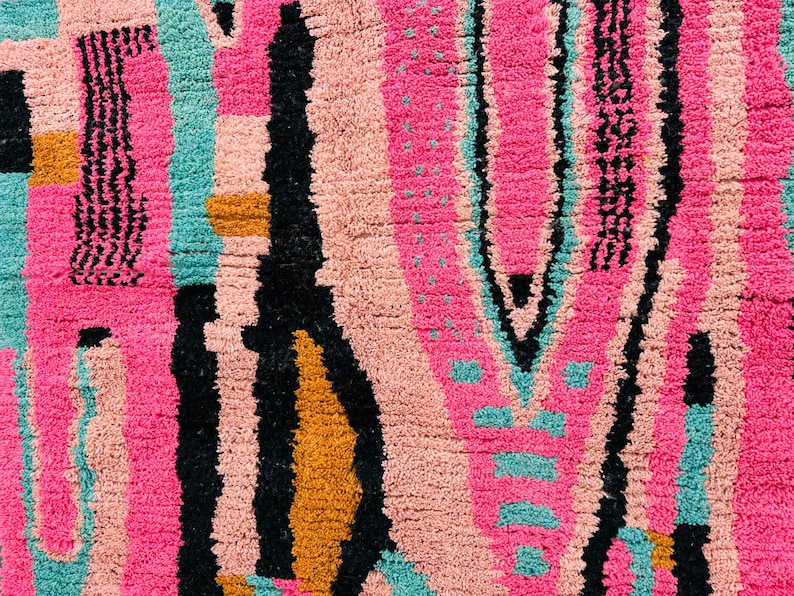 May include: Close-up of a colorful rug with a geometric pattern. The rug features a vibrant mix of colors, including pink, black, teal, and gold. The design includes vertical stripes and abstract shapes, creating a textured surface.