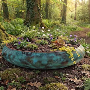 Puede incluir: Una maceta baja, redonda y con forma de cuenco con un acabado de pátina desgastada en tonos verde azulado y marrón. La maceta está llena de tierra oscura, follaje verde, pequeñas flores blancas y flores moradas, creando un contraste vibrante.