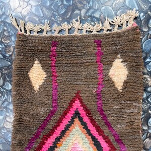 May include: A brown, pink, and white Moroccan rug with a diamond pattern and fringe. The rug is made of wool and has a hand-woven texture.