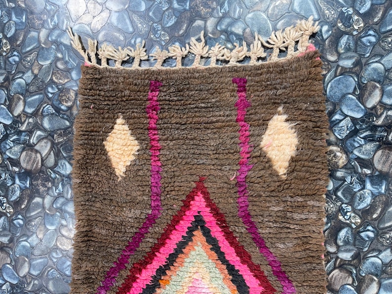 May include: A brown, pink, and white Moroccan rug with a diamond pattern and fringe. The rug is made of wool and has a hand-woven texture.