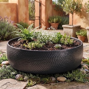 Pode incluir: Um vaso grande, redondo e cinza-escuro com um acabamento de metal martelado e texturizado. O vaso está cheio de terra escura e uma variedade de pequenas plantas verdes, incluindo fetos e suculentas, sobre um pátio de pedra.